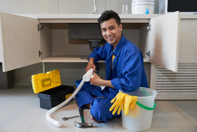 cheerful asian plumber sitting floor repairing kitchen sink 1